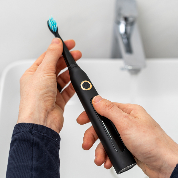 Twee handen houden een zwarte elektrische tandenborstel vast voor een wastafel en kraan in de badkamer.