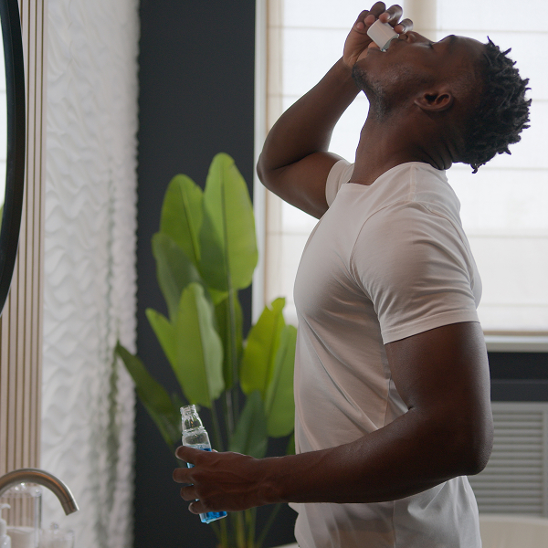 Une personne utilise un bain de bouche dans une salle de bain tout en tenant un flacon de solution buccale devant un miroir.