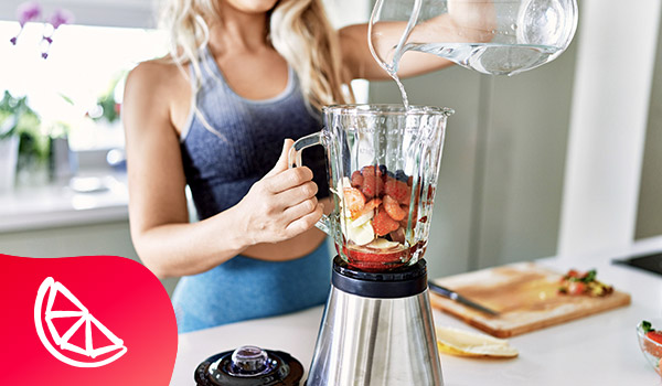 Een vrouw in sportieve kledij voegt water toe aan een blender waarvan de kom is gevuld met stukjes banaan, stukjes aardbei, frambozen en blauwbessen.