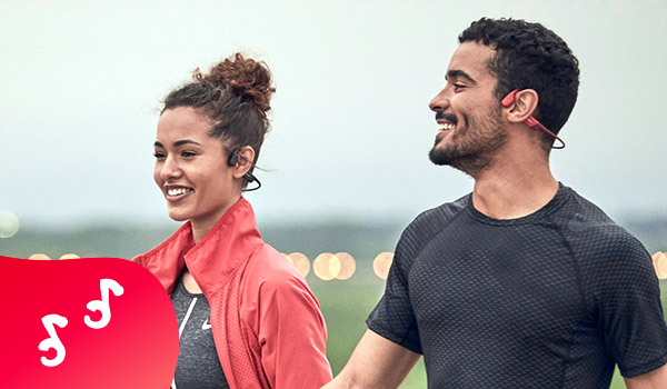 Een man en een vrouw in sportieve kledij wandelen glimlachend. Ze dragen beiden sportoortjes, zwarte voor haar en roze voor hem.