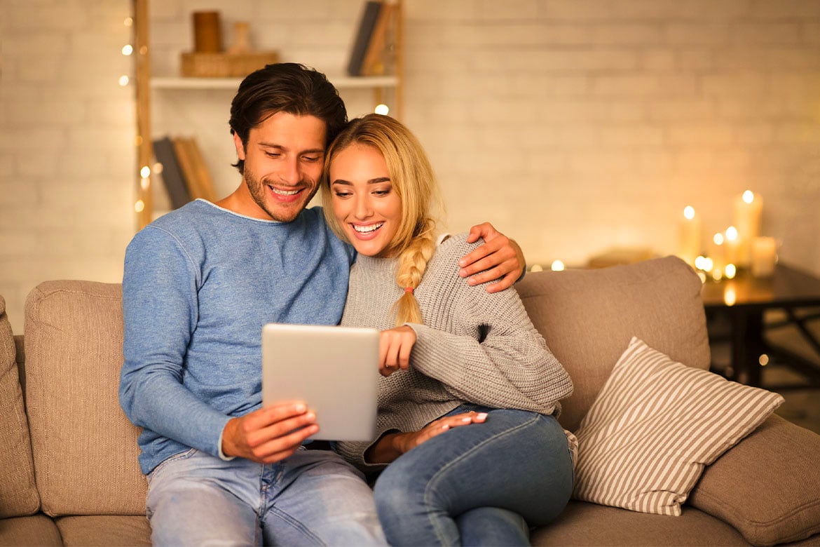 Een koppel, vrouw en man, zittend in de zetel, kijken samen naar een tablet. De man heeft zijn arm rond de vrouw.