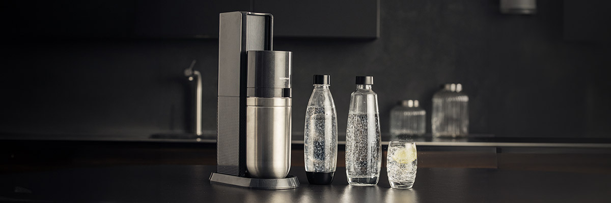 Un appareil Sodastream Duo Black, les deux bouteilles et un verre remplis d'eau gazeuse, dans une cuisine sombre