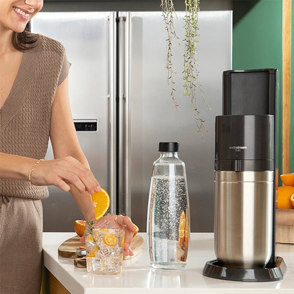 Une dame qui met une tranche d'orange dans un verre d'eau gazeuse,  ct de l'appareil Sodastream Duo Black