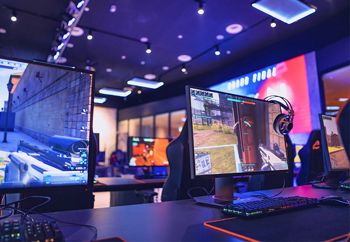Rang�e de postes de gaming avec des �crans affichant une sc�ne de jeu vid�o en vue subjective et des casques suspendus, dans une salle �clair�e par des lumi�res color�es