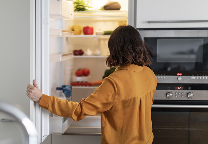 Vue de dos d�une personne ouvrant la porte d�un r�frig�rateur rempli d�aliments dans une cuisine avec un four et un micro‑ondes int�gr�s