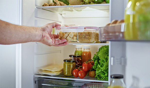 Main d�posant un r�cipient transparent contenant des aliments dans un r�frig�rateur ouvert rempli de l�gumes, de bouteilles et de bocaux