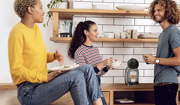 Trois personnes discutant dans une cuisine avec des �tag�res en bois, de la vaisselle visible et une machine � caf� pos�e sur le plan de travail.