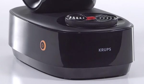 Gros plan sur la base d�une machine � caf� noire de la marque Krups, montrant le bouton d�alimentation et une grille circulaire sur le dessus.