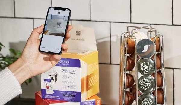 Main tenant un smartphone scannant un QR code sur une bo�te pos�e sur un plan de travail, avec un porte‑capsules contenant des dosettes Dolce Gusto � droite.