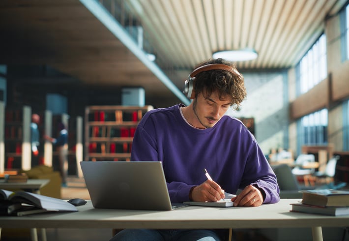 Persoon gebruikt een laptop en maakt notities in een bibliotheek.
